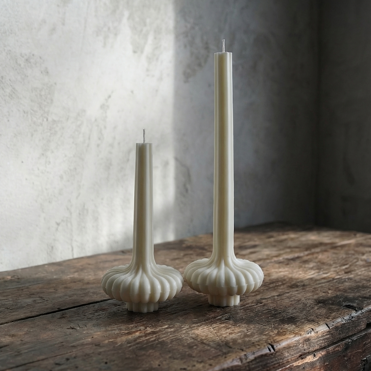 Two white candle holders with candles on a rustic wooden surface against a textured wall.