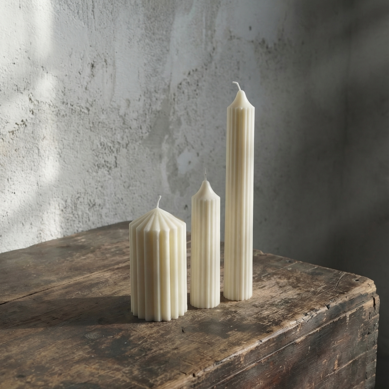 Three white candles of different shapes on a wooden surface with a textured wall background
