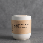 White candle in a glass container with a brown label on a gray background